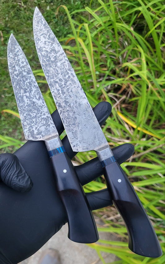 Two kitchen knives with patterned steel blades and black handles are held by a gloved hand outdoors in front of green grass.